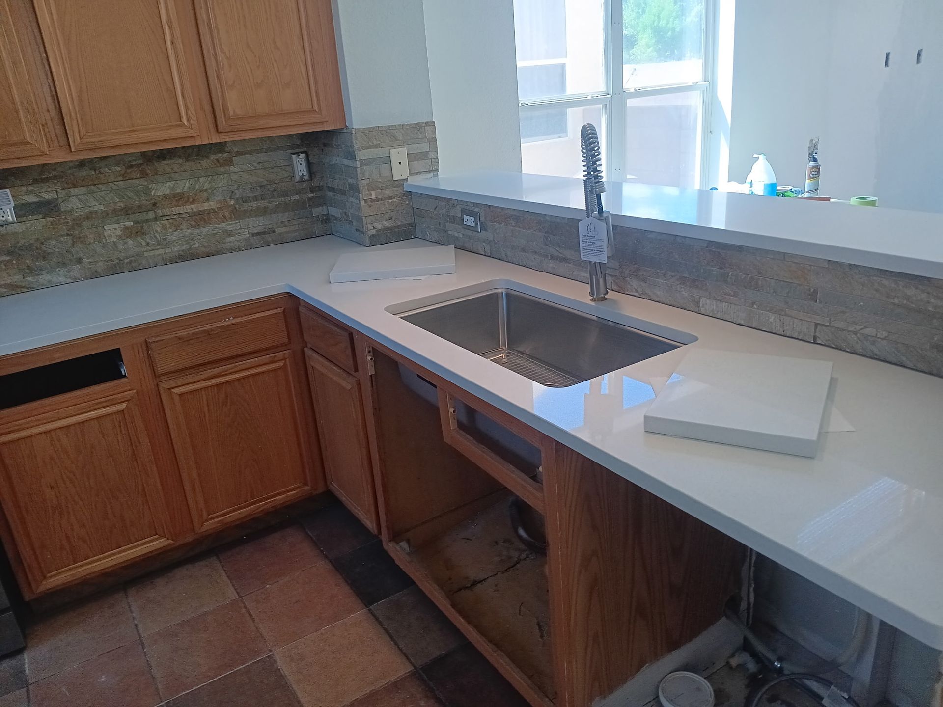 Kitchen with light-colored countertops, cabinets, stone backsplash, and a stainless steel sink near a window.