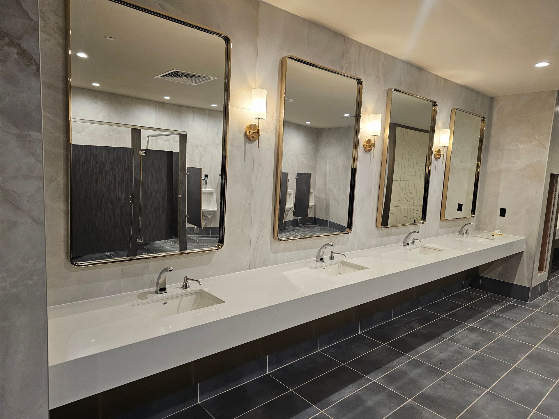 Public restroom with three framed mirrors, sinks, and stalls. Neutral colors, tiled floors.