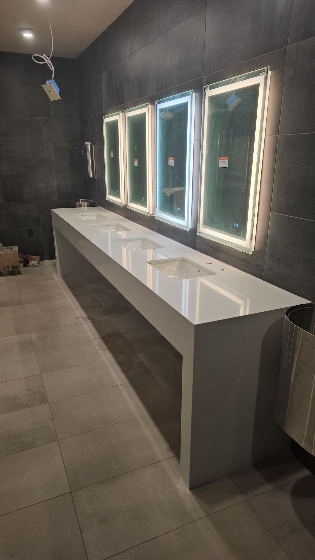 Long white bathroom vanity with sinks and mirrors, dark grey wall, and gray floor.