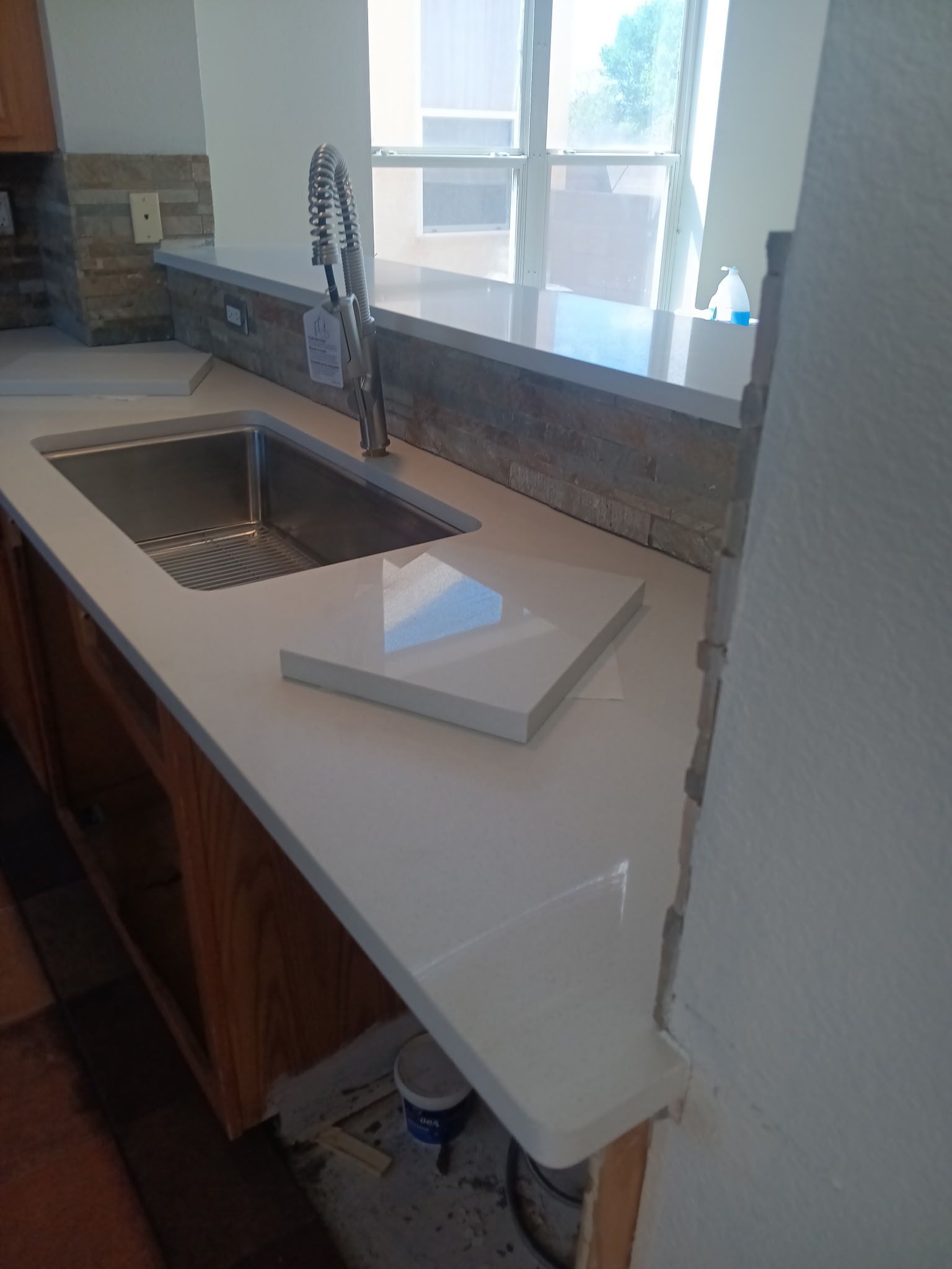 Kitchen countertop with sink, faucet, and backsplash. White countertop, wooden cabinets, and a view of outside.