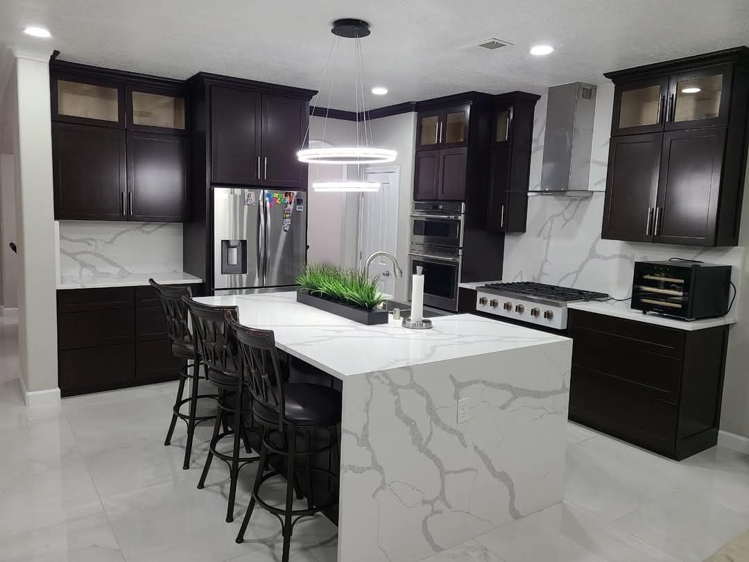 Dark-cabinet kitchen with white marble countertops and island, stainless steel appliances, and a modern light fixture.