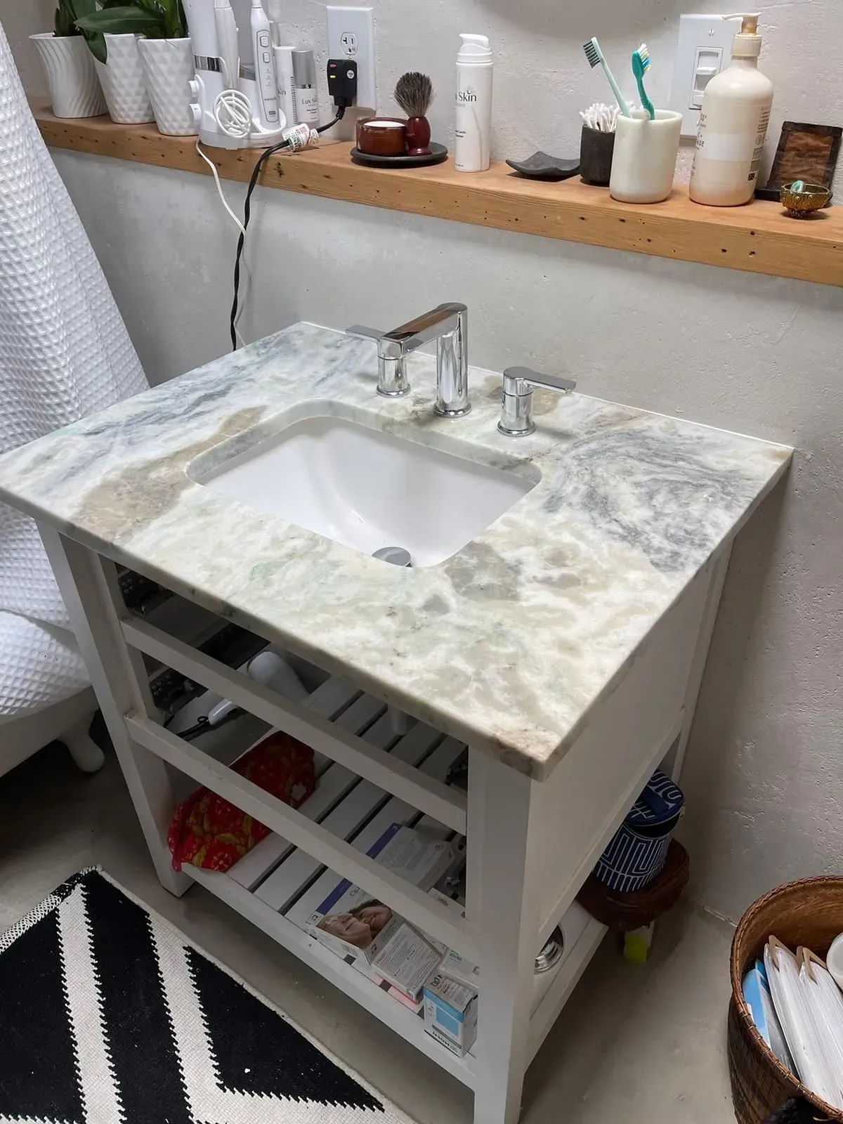 White marble bathroom vanity with sink, open shelves, and toiletries on a wooden shelf above.