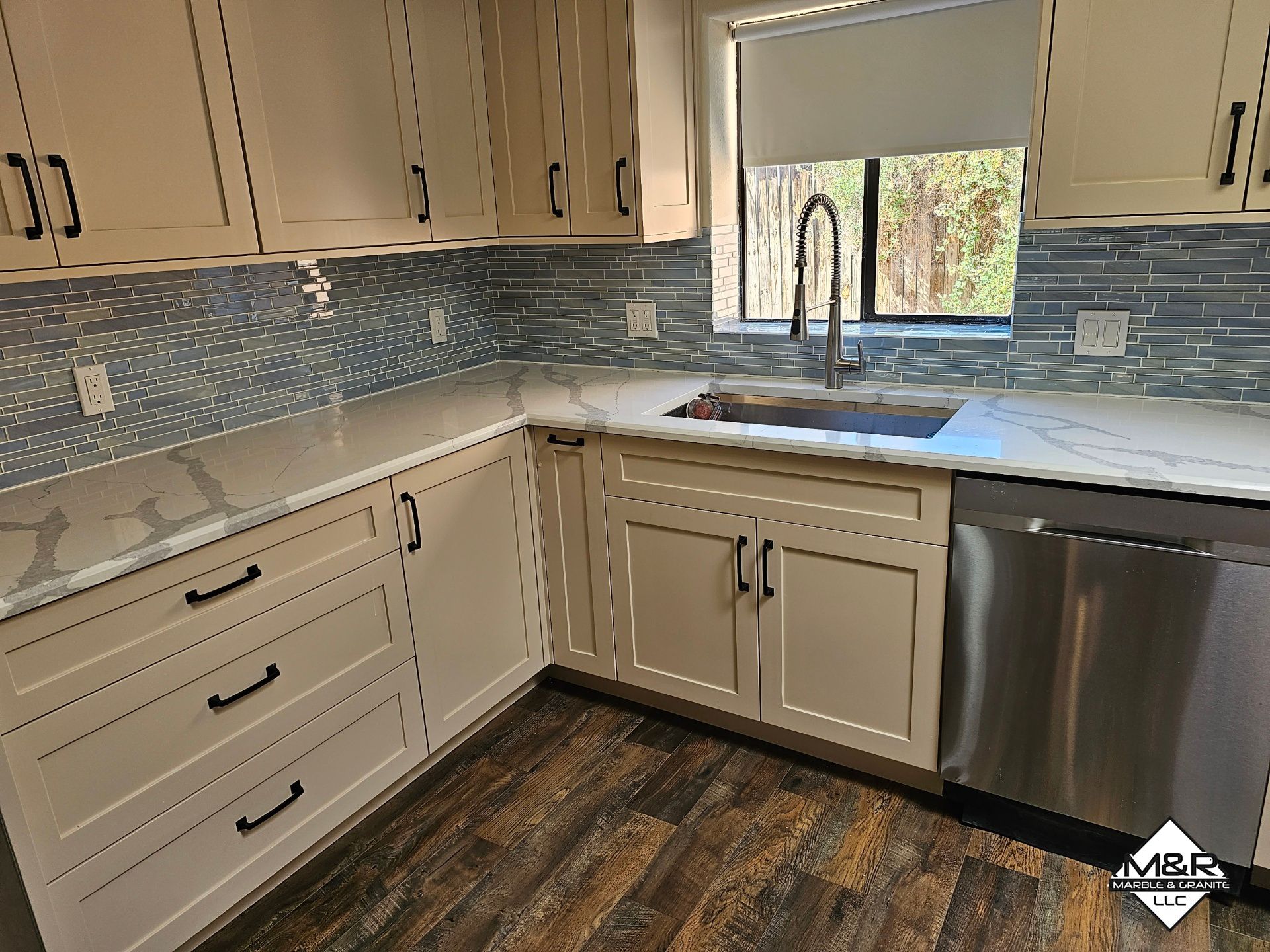 A modern kitchen with off-white cabinets, marble countertops, blue tiled backsplash, and stainless steel appliances.