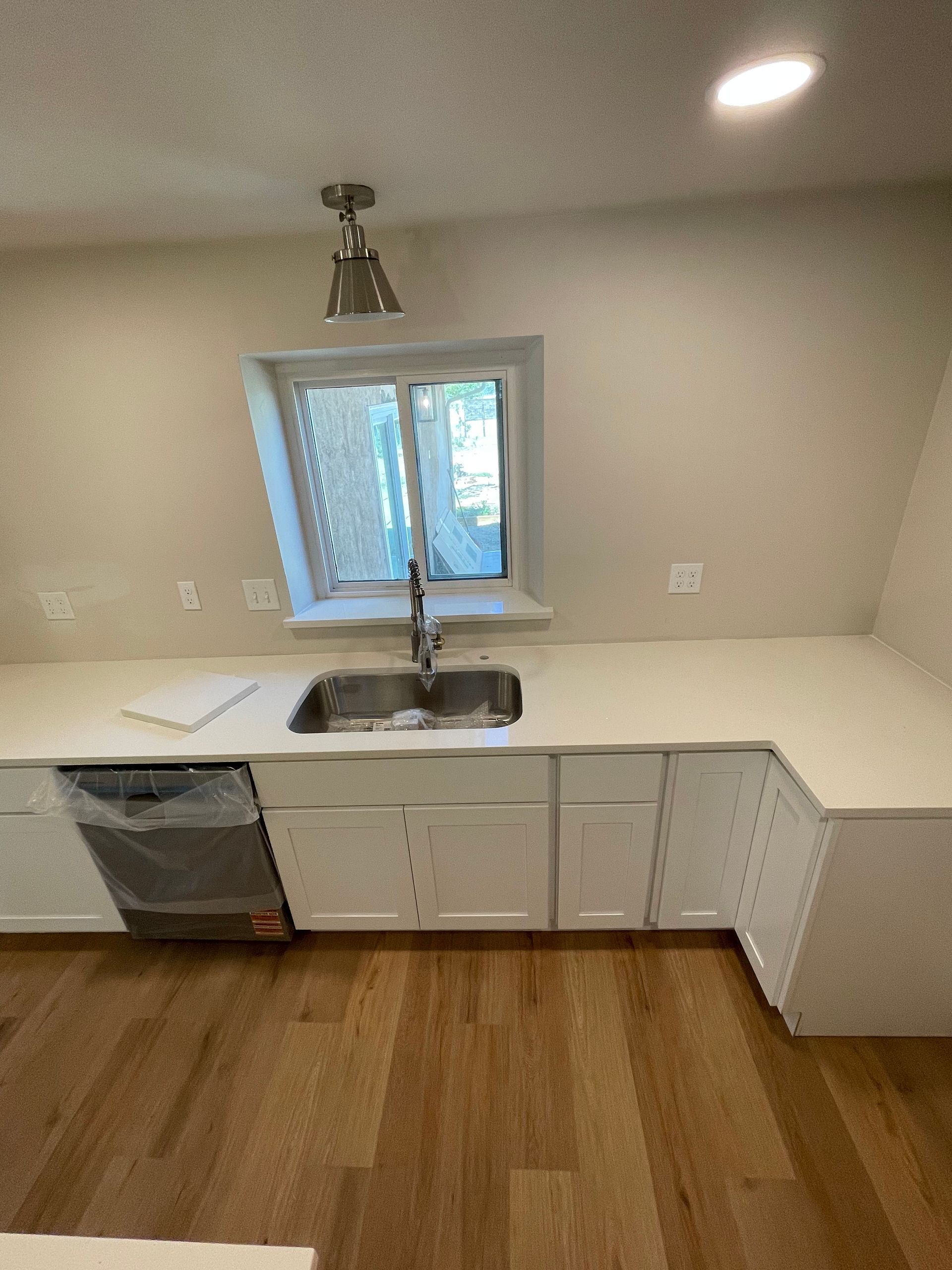 White kitchen with stainless steel sink, dishwasher, and cabinetry, with a window above and hardwood floors.