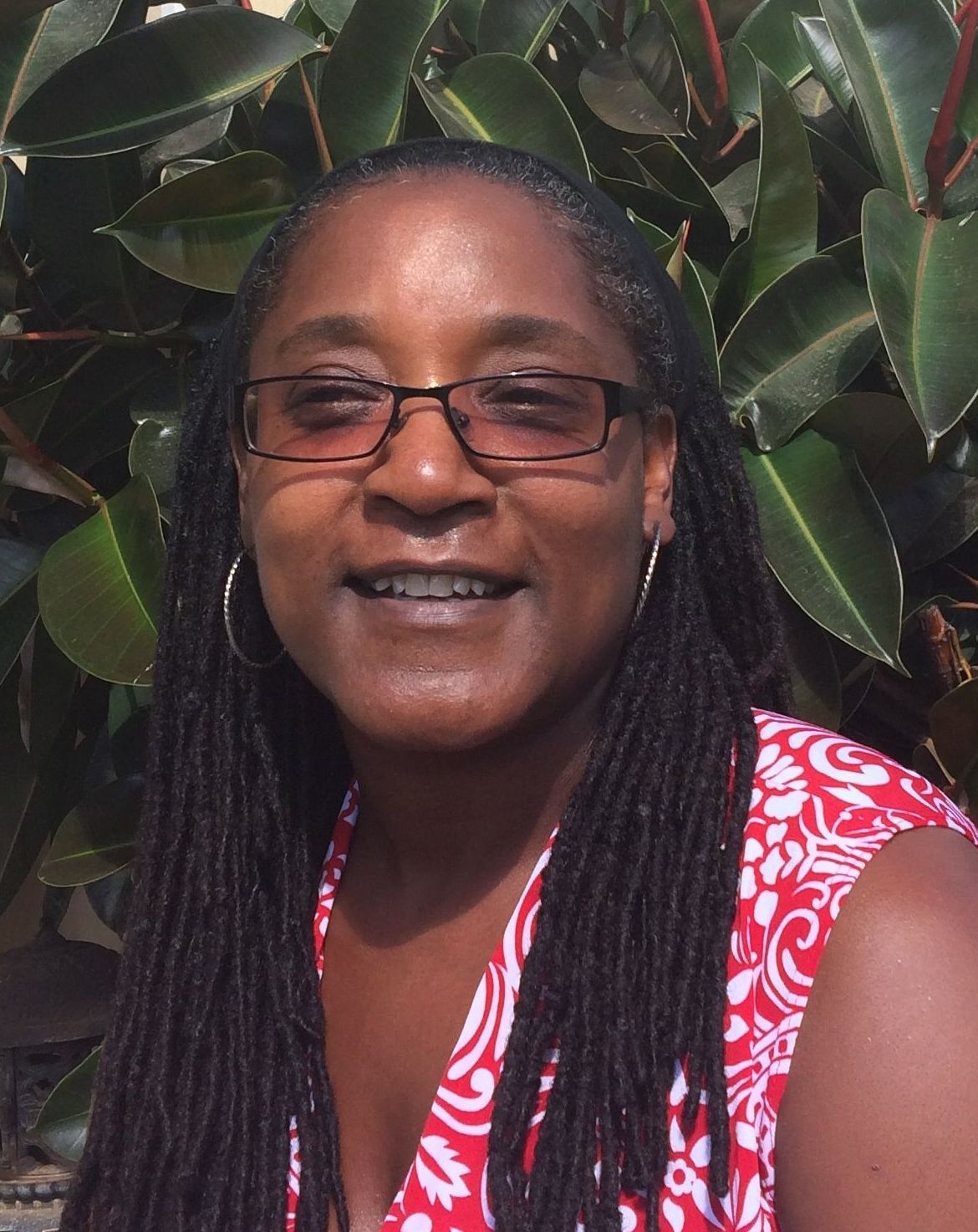 A woman wearing glasses and dreadlocks is smiling in front of a plant.