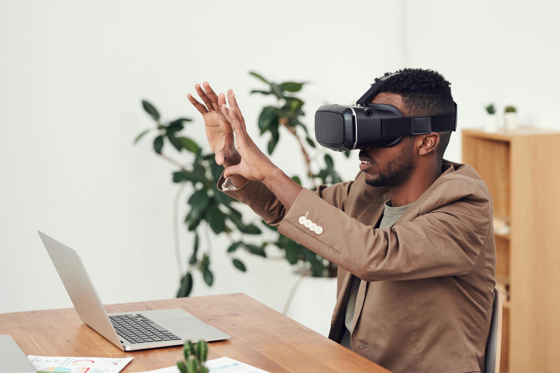 A man wearing a virtual reality headset is sitting at a desk with a laptop.