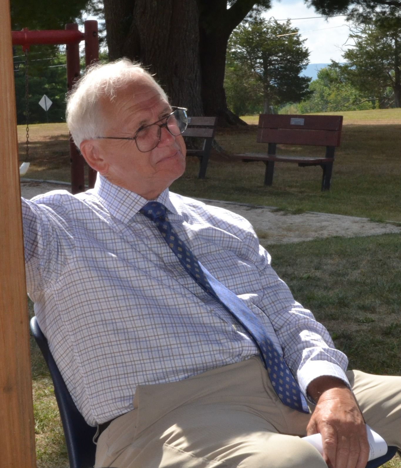 Older man, light skin, wearing glasses, blue tie, and checked shirt, sits outside in park, looking off.