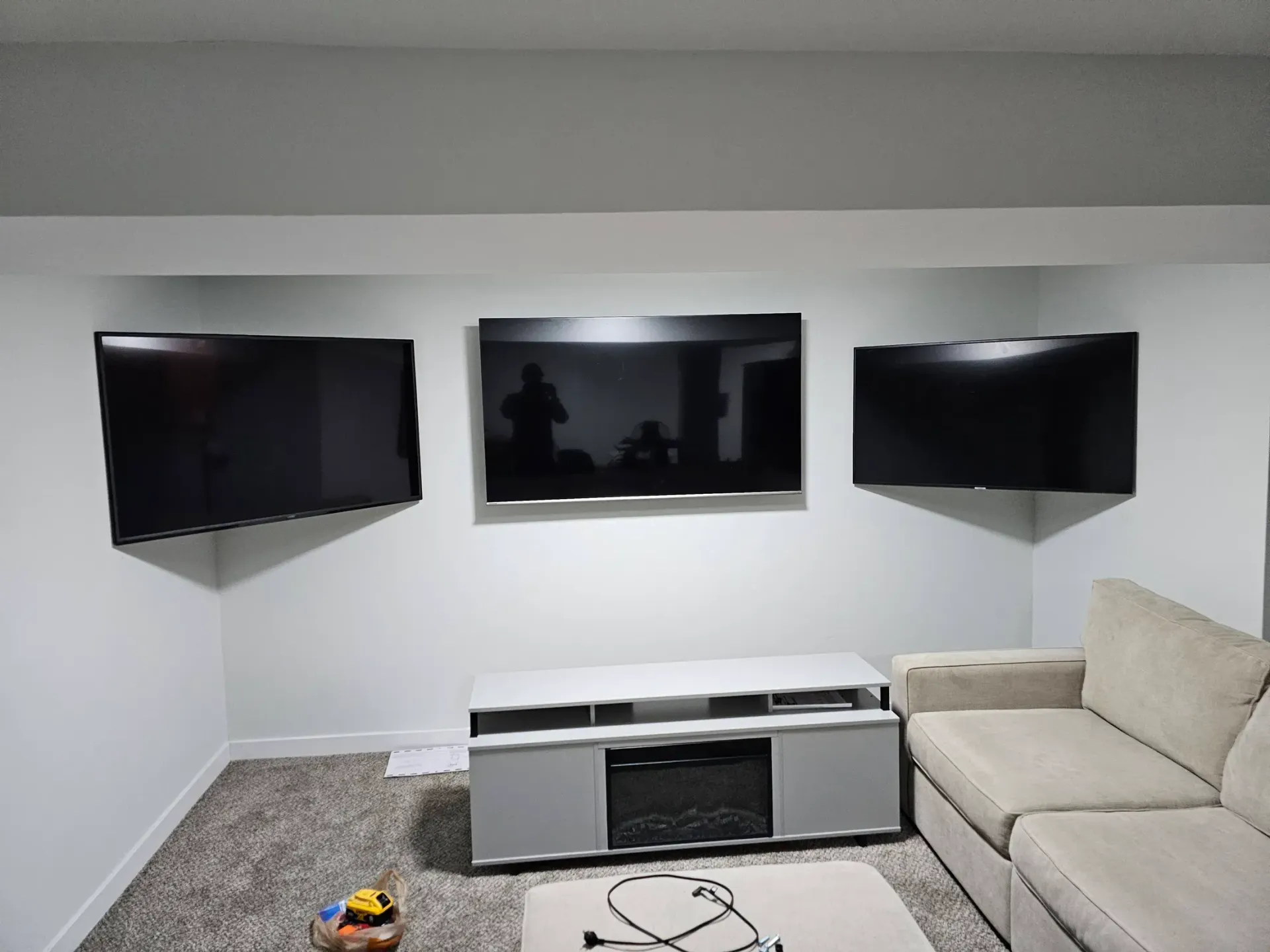 Three wall-mounted TVs above a gray media console, with a beige sofa on the right and carpeted flooring.
