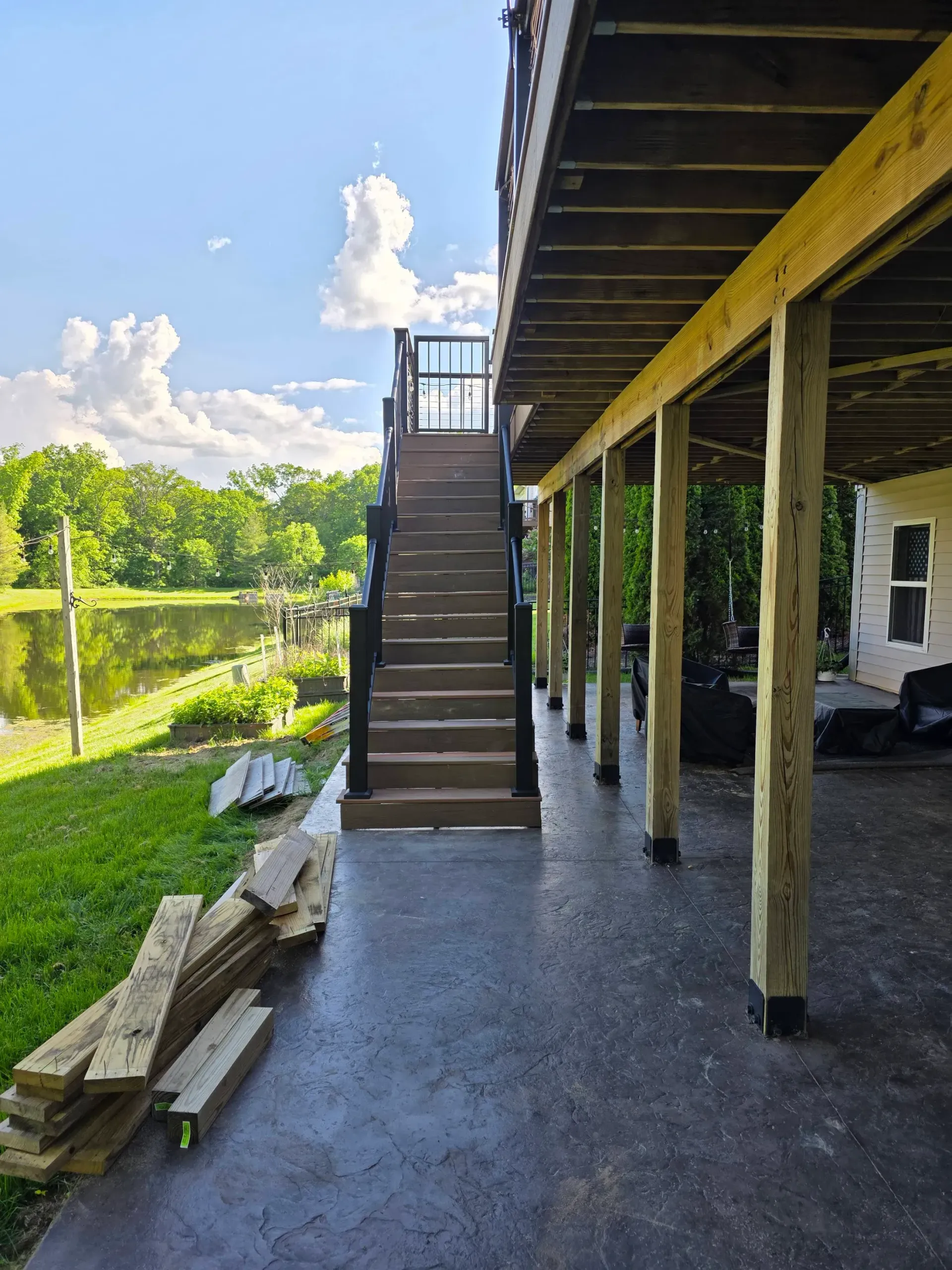 Deck with stairs leading up, supported by wooden posts. Concrete patio area, lake visible in the background.