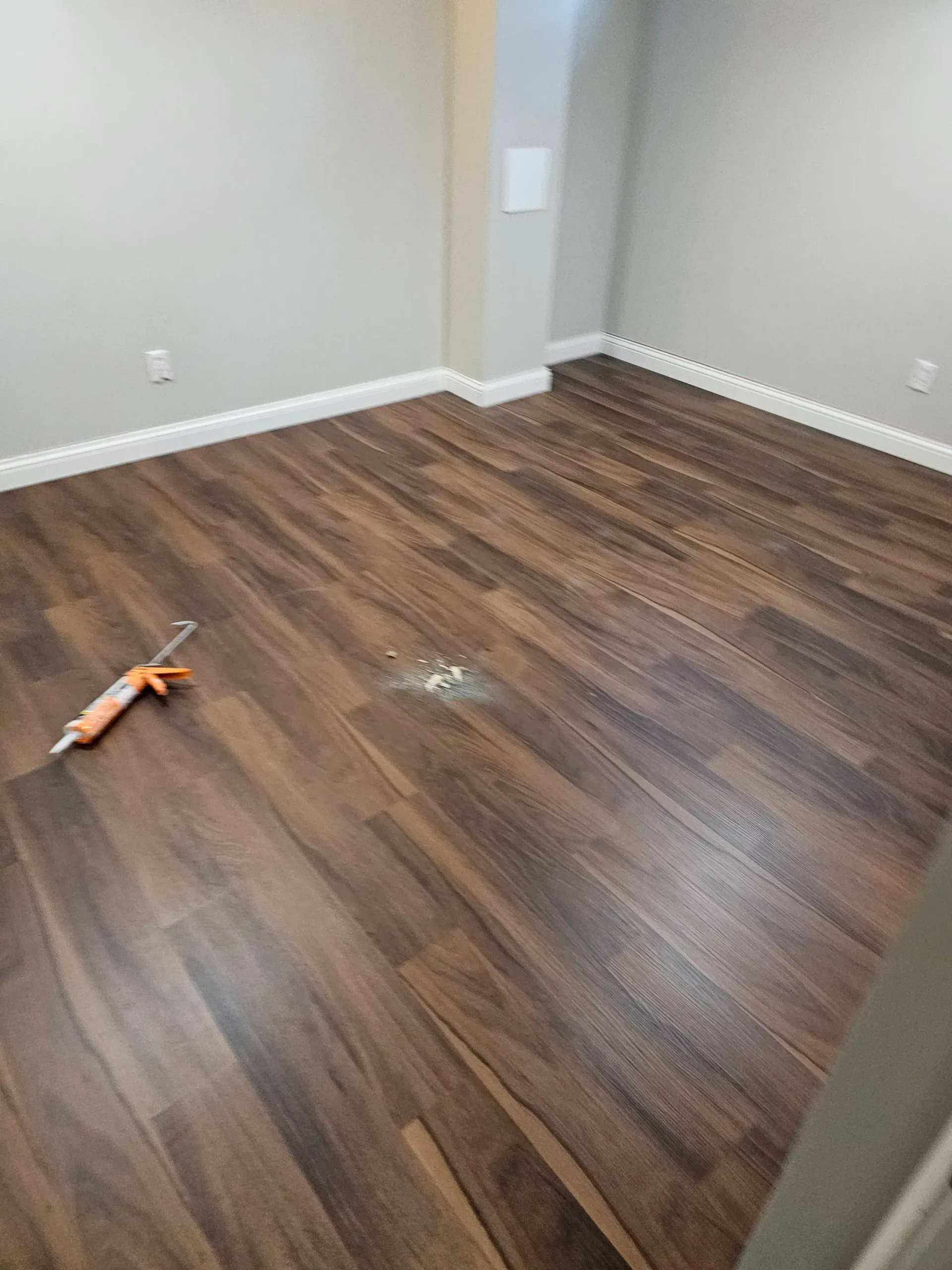 Room with wood-look flooring and light gray walls. Caulk gun and spilled caulk on the floor.