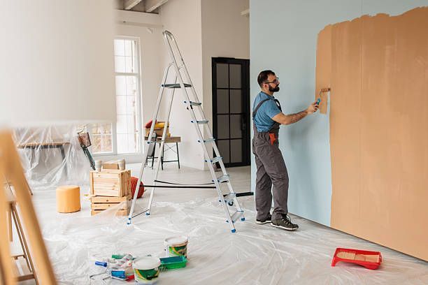 Man painting a wall blue with a roller, standing on a step ladder inside a room.