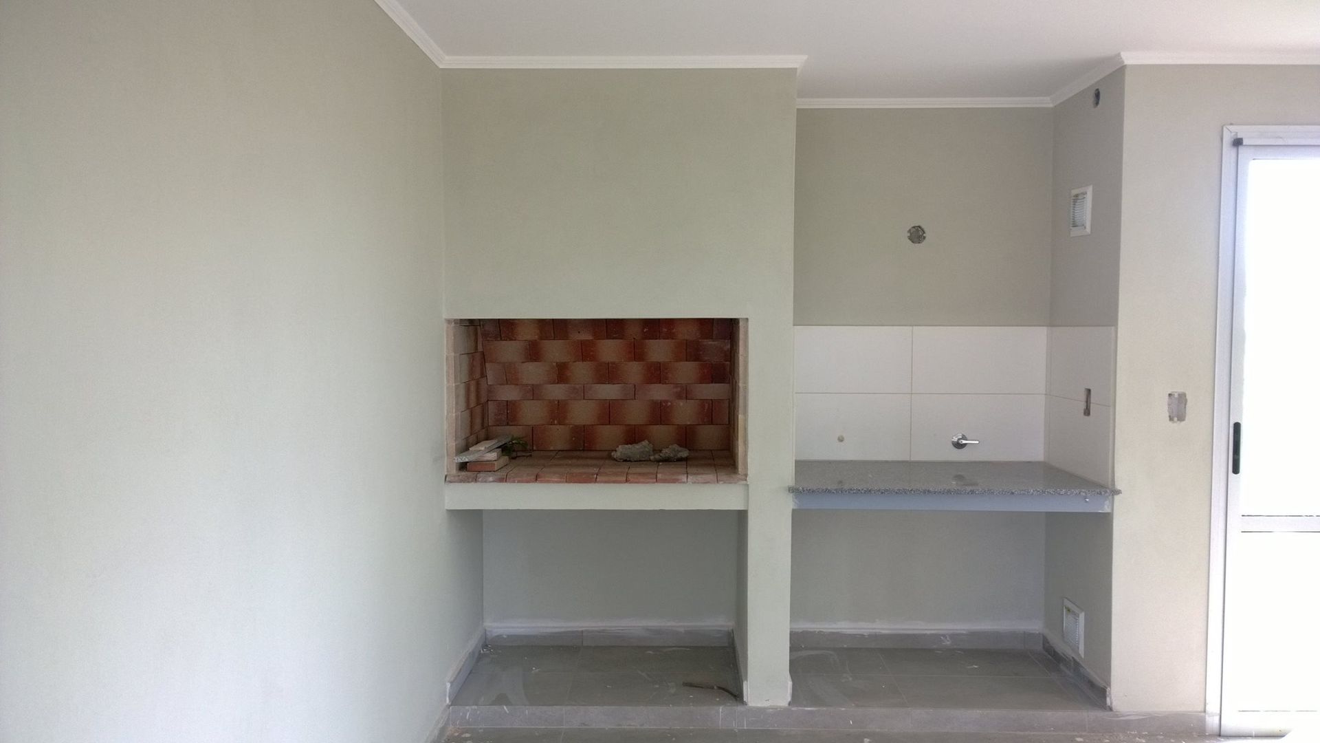 Habitación pequeña sin terminar con un escritorio blanco, una estantería empotrada y una puerta corredera de cristal.