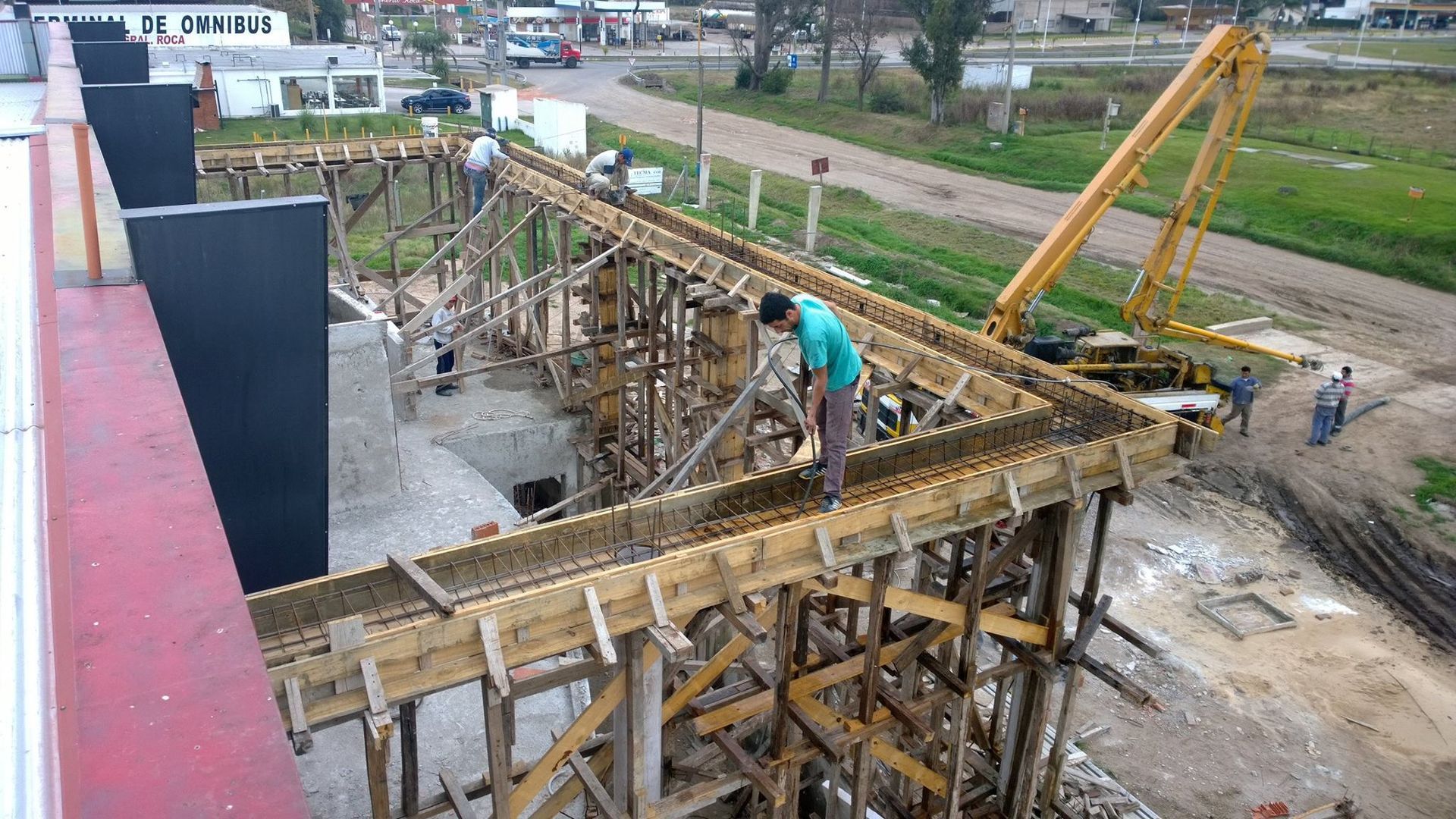Obreros de la construcción en andamios de madera junto a un canal, con una grúa amarilla y encofrados de hormigón.