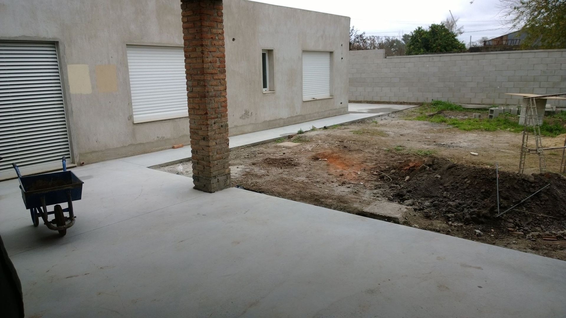 Patio de concreto vacío junto a una casa beige, con un pequeño carrito y un patio desnudo bajo cielos nublados.