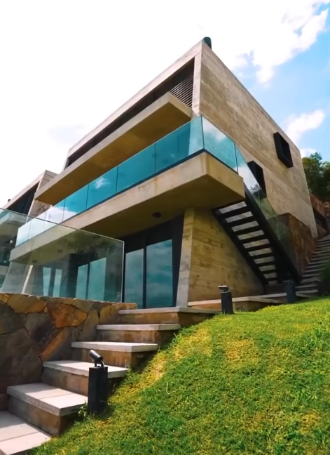 Casa moderna con balcones de cristal, detalles de piedra y escaleras que suben por una ladera cubierta de hierba.