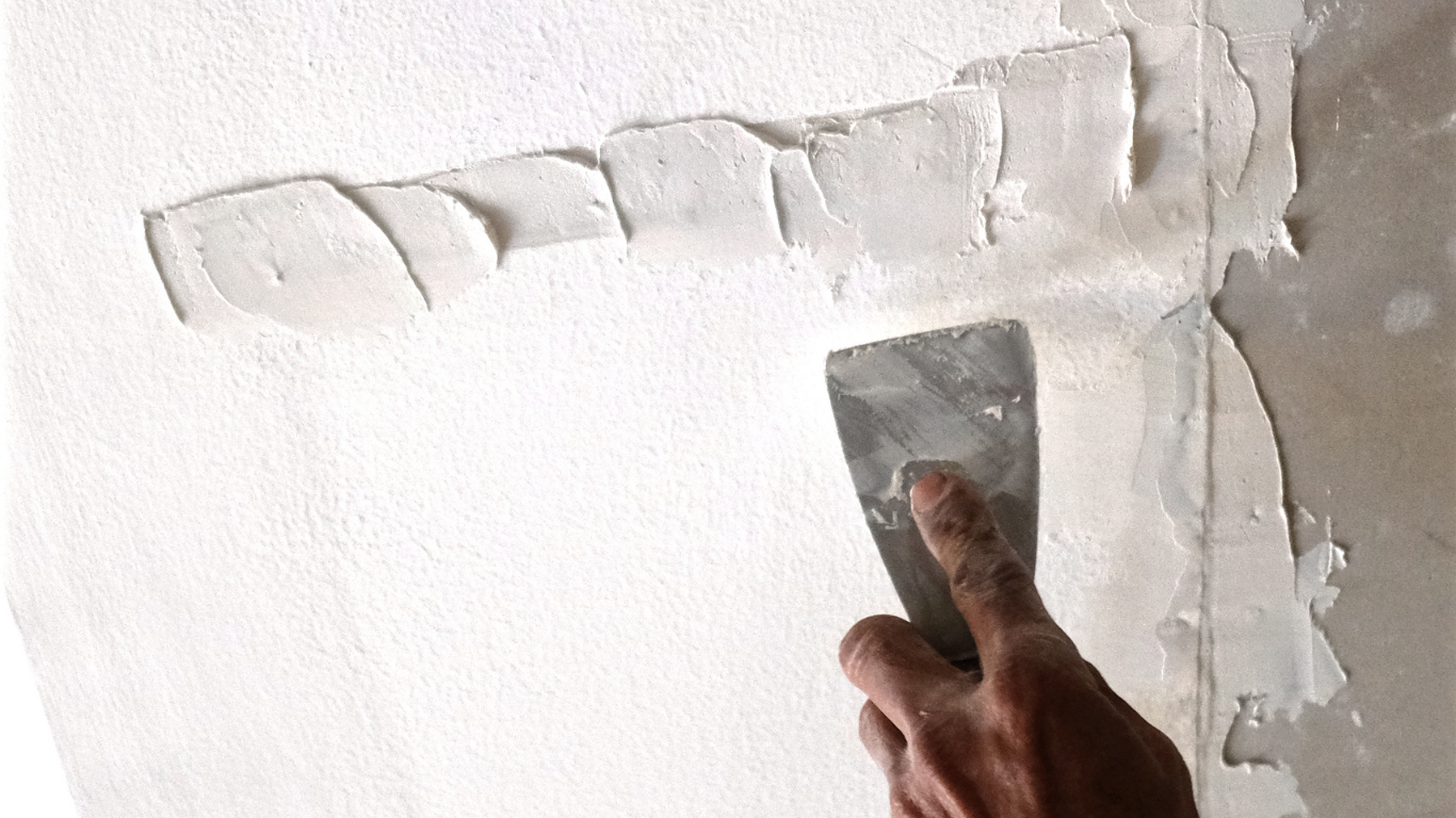 Hand using a putty knife to spread white joint compound on a wall.