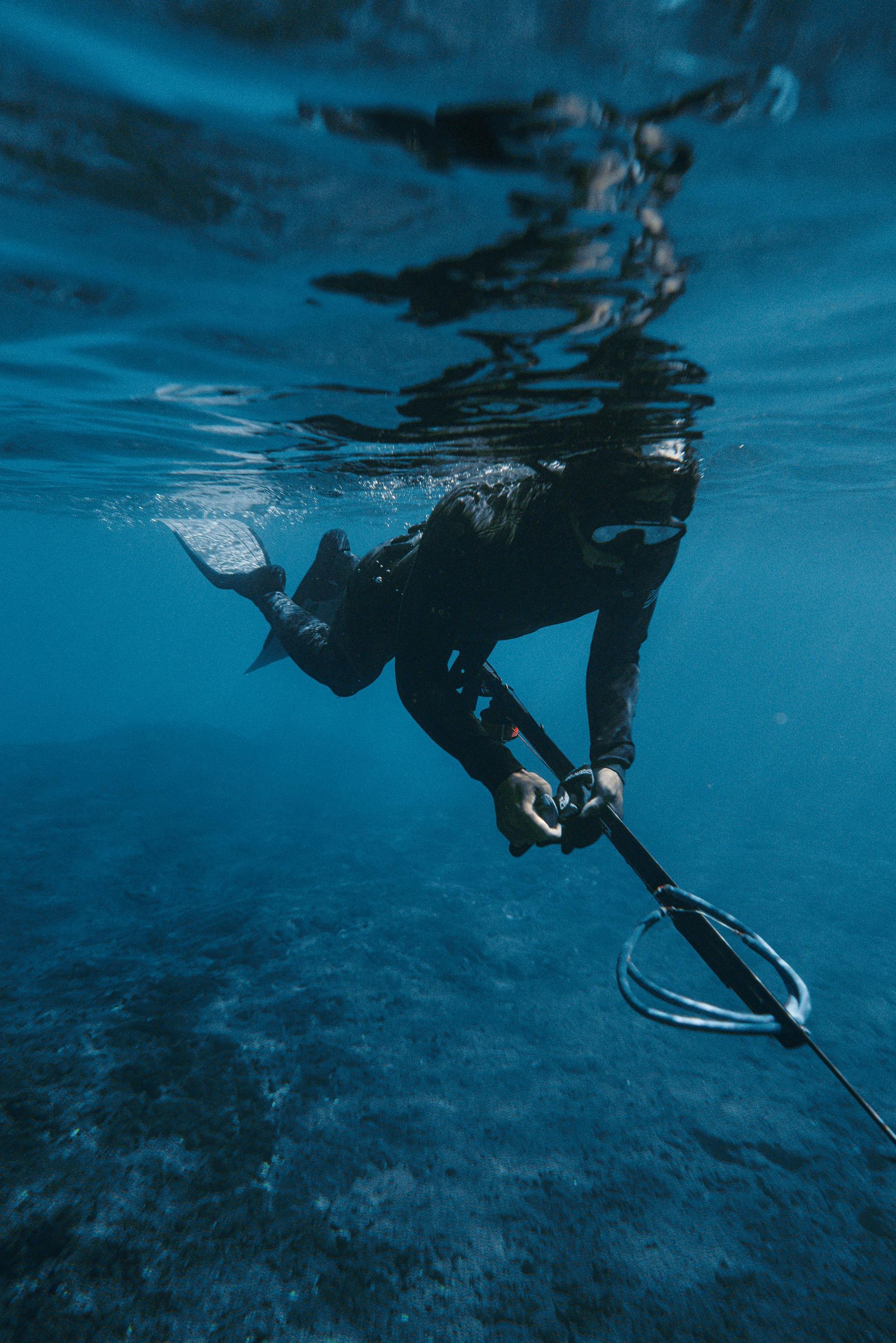 Person spearfishing underwater in blue water, holding spear gun, with flippers.