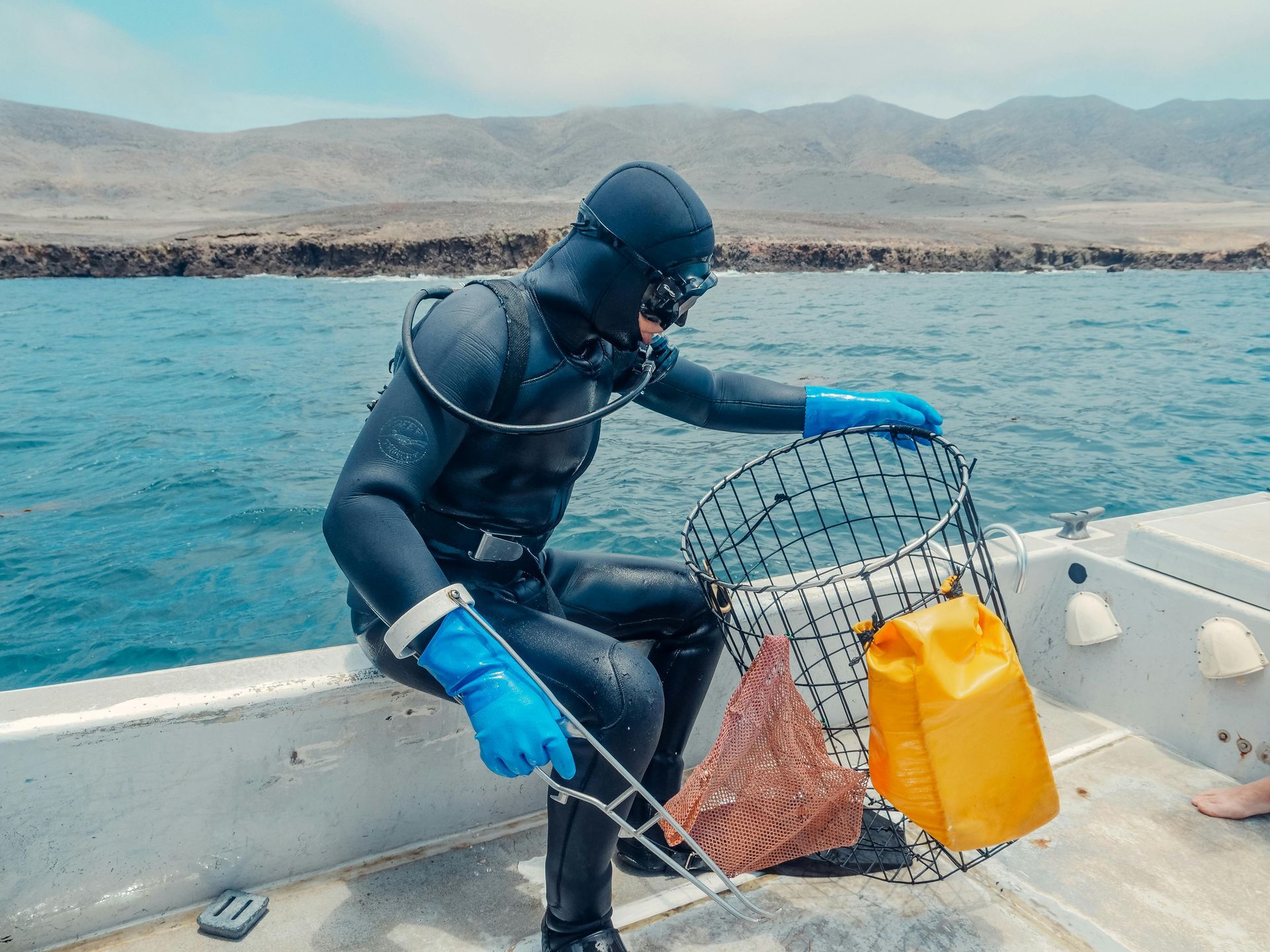 A diver on a boat hauls a fishing trap from blue water with mountains in the background