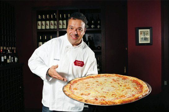 Chef holding a large cheese pizza, smiling, in a restaurant with wine bottles on shelves behind him.