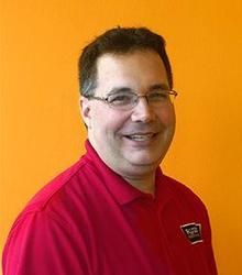 Man with glasses and a red polo shirt smiles at the camera, orange background.