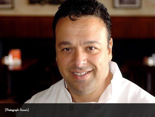 Smiling man in a white chef's coat; dark hair and skin; interior restaurant setting.