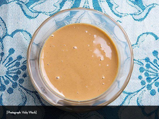 A small glass bowl holding a smooth, tan-colored sauce, set on a blue floral patterned tablecloth.