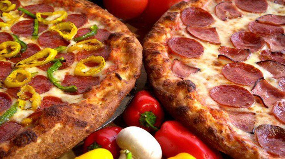 Two pizzas, one with peppers and the other with pepperoni, surrounded by vegetables.
