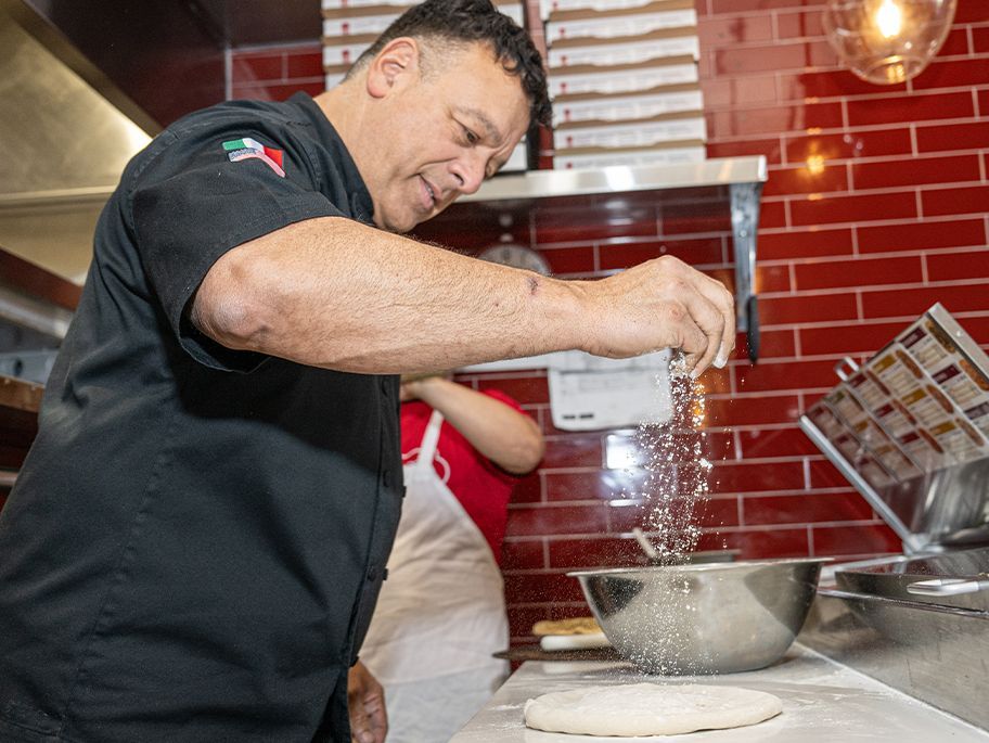 Chef sprinkling flour on pizza dough in a kitchen with red brick walls.