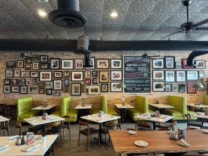 Interior of a restaurant with lime green booths, square tables, and a brick wall decorated with framed photos and a menu board.