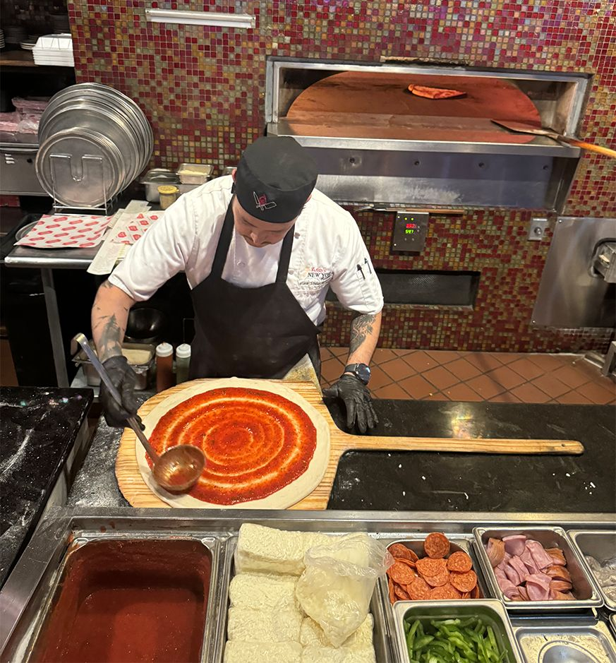 Chef in black apron preparing pizza, spreading sauce on the dough with wooden paddle, next to an oven.