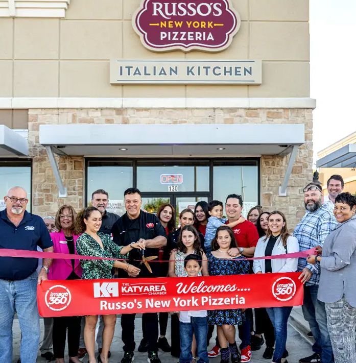 People cutting a ribbon at the grand opening of Russo's New York Pizzeria in Katy, Texas. A man in black cuts the ribbon, surrounded by a crowd.