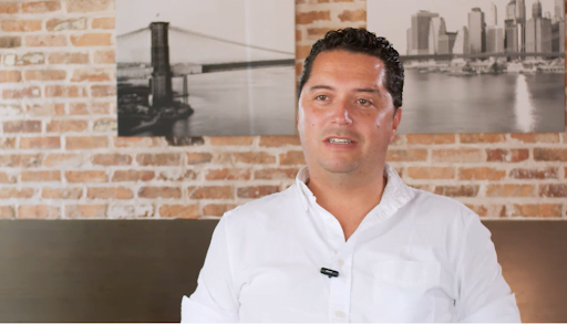 Man in white shirt speaking, in front of a brick wall and a cityscape photo.