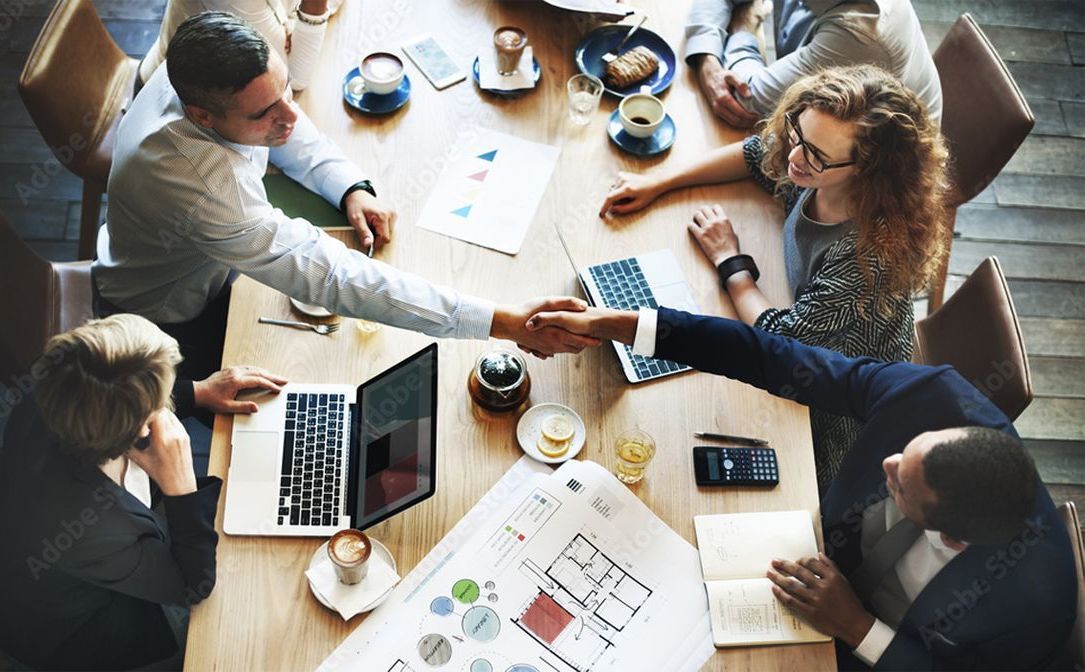 Business people shaking hands at a table, surrounded by documents, laptops, and coffee cups. Top-down view in a well-lit meeting room.