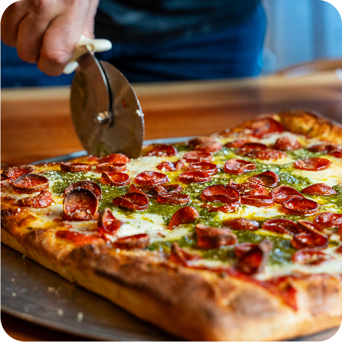 Person slicing a rectangular pizza topped with pepperoni and green sauce on a metal tray.