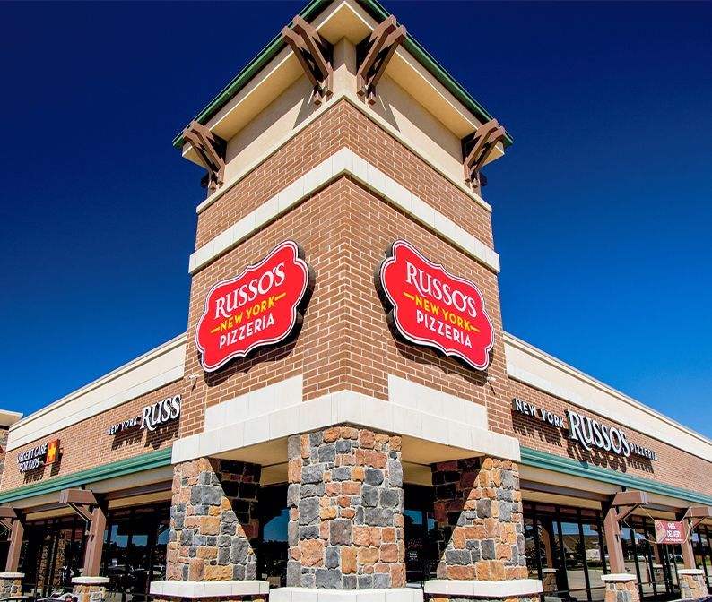Russo's New York Pizzeria storefront with brick and stone exterior, red signs, and a clear blue sky.