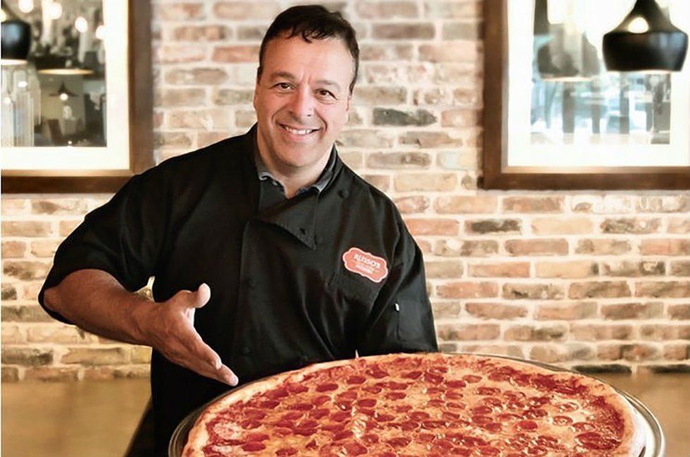Chef smiling, presenting a large pepperoni pizza in a restaurant setting with a brick wall background.