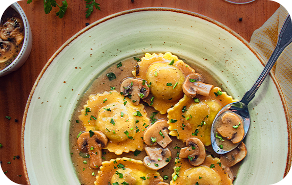 Ravioli in mushroom sauce served in a rustic plate, garnished with herbs. A spoonful of sauce with mushrooms and pasta is in the foreground.