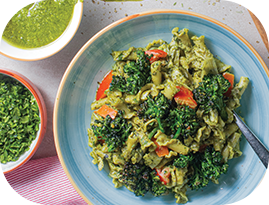 Pasta with pesto sauce, kale, and tomatoes in a blue bowl, with a bowl of pesto sauce and chopped greens nearby.