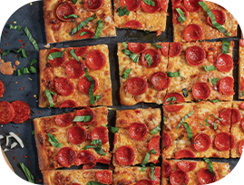 Square-cut pepperoni pizza with basil on a dark surface. Slices of pizza are close together, with visible toppings.