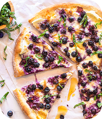 Blueberry pizza with cheese, basil, and drizzle, sliced on parchment paper.