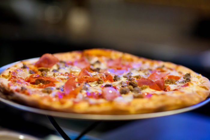 Close-up of a pizza on a plate, with toppings including meat and vegetables. The pizza sits in a dimly lit environment.