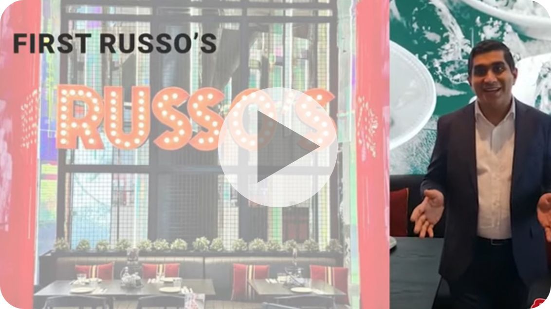 Man standing in a restaurant, gesturing. "First Russo's" sign behind him.