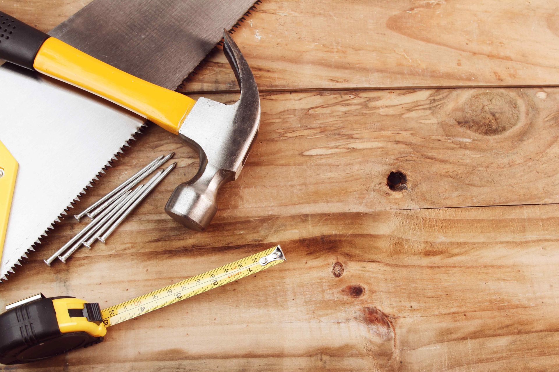 A hammer , saw , tape measure , and nails are on a wooden table.