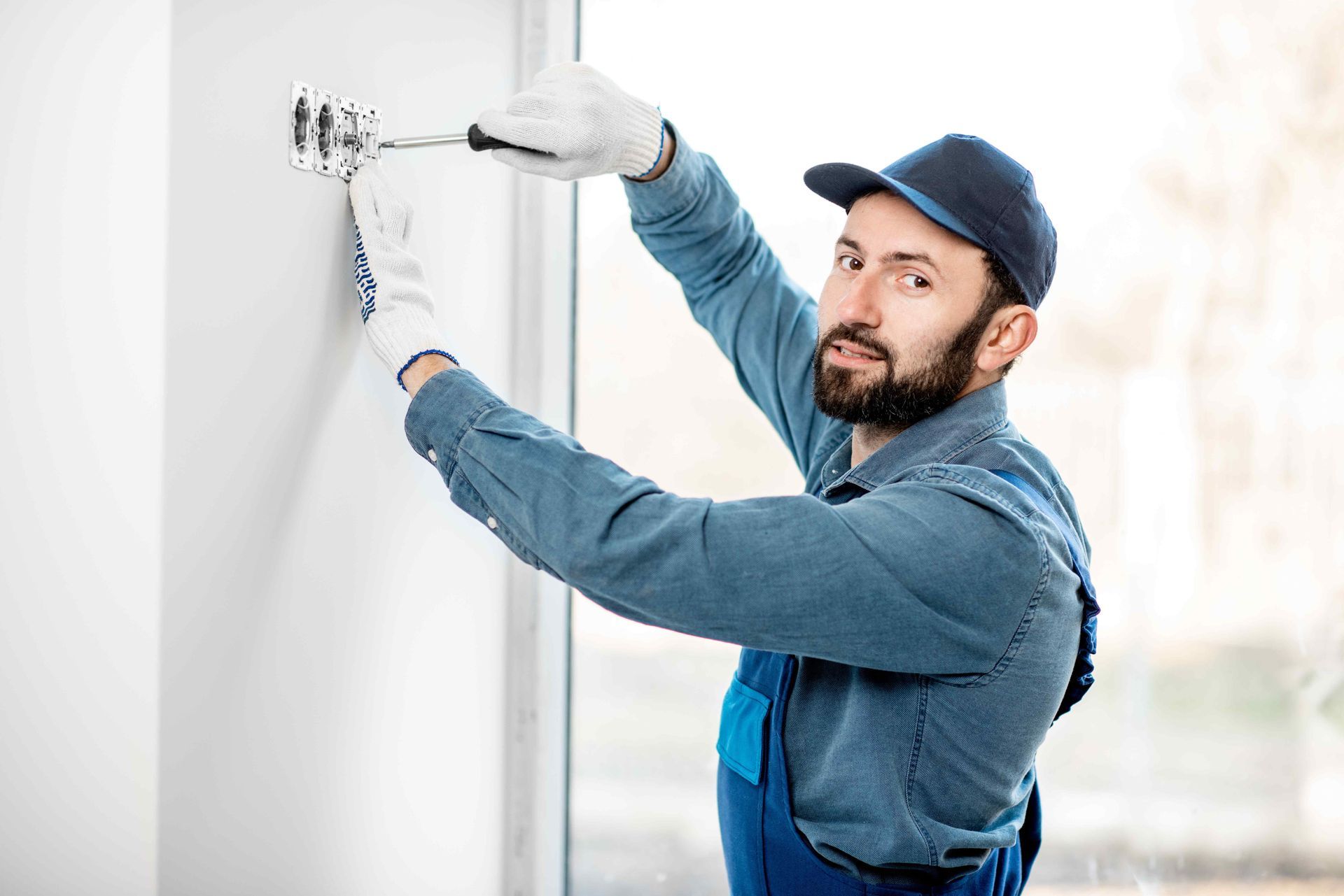 Electrician in blue uniform, cap, and gloves, tightening a wall outlet with a screwdriver.
