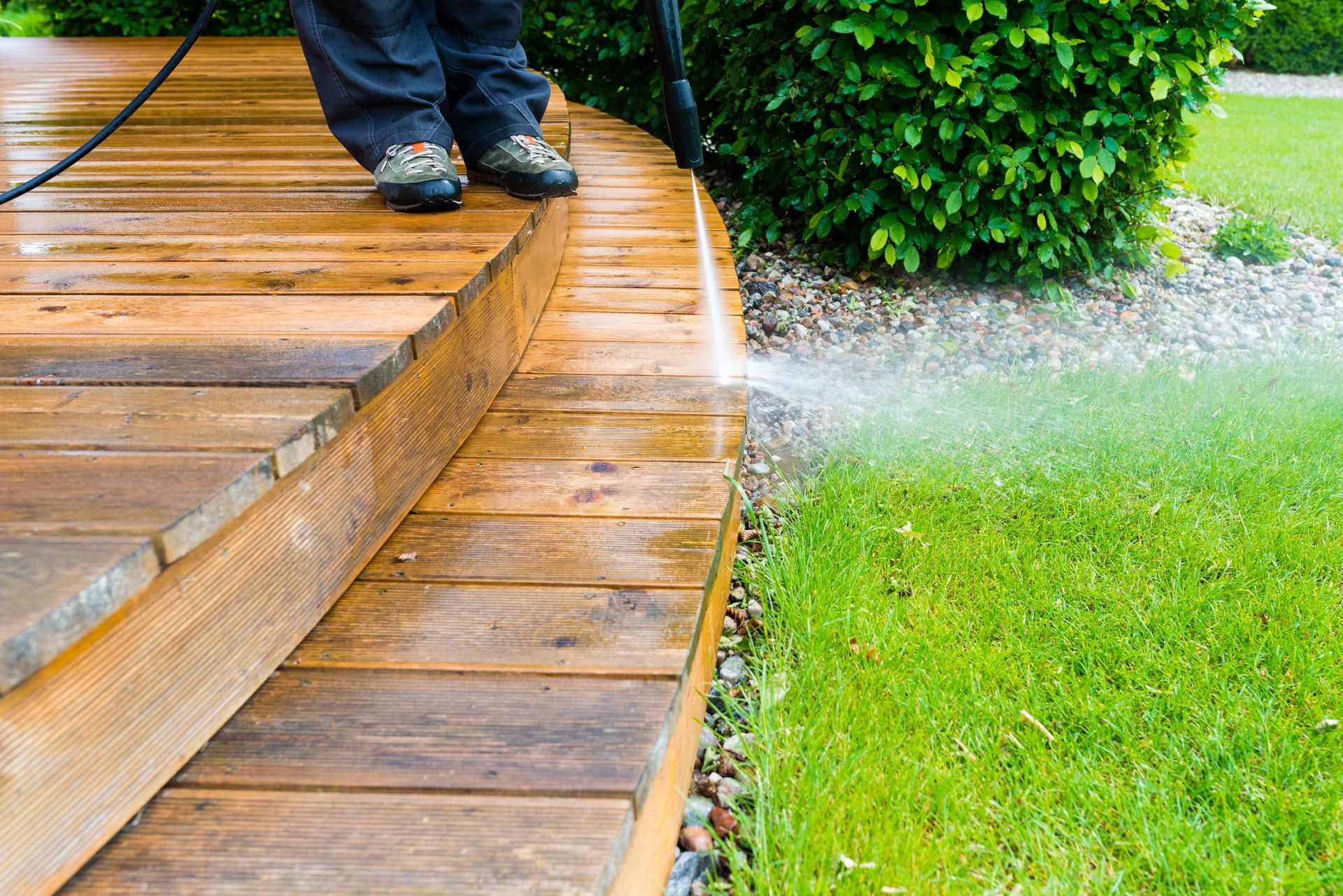 Person power washing a wooden deck next to a green lawn and bushes.