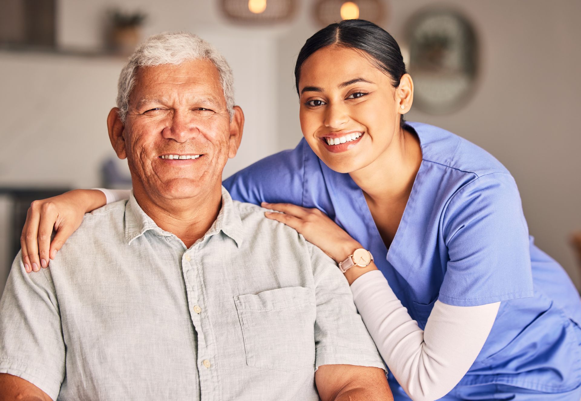 Elderly man seated with nurse in blue scrubs, hand on shoulder, both facing camera.