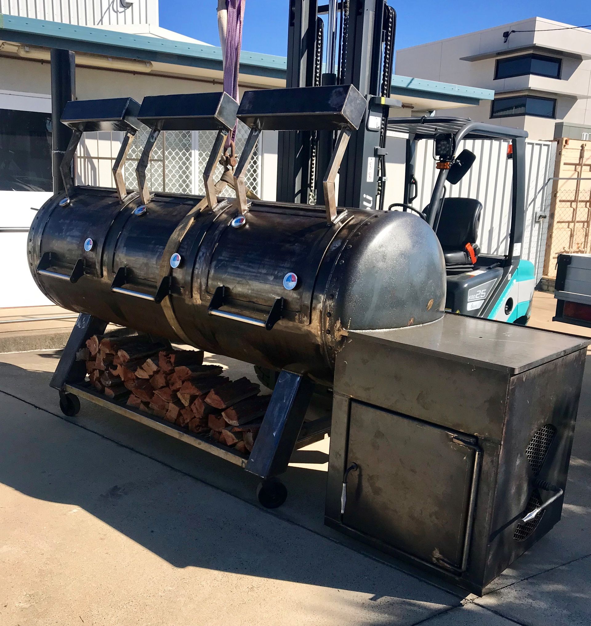 Forklift lifting a large, black smoker grill with a wood storage rack — Townsville Stainless Steel in Garbutt, QLD