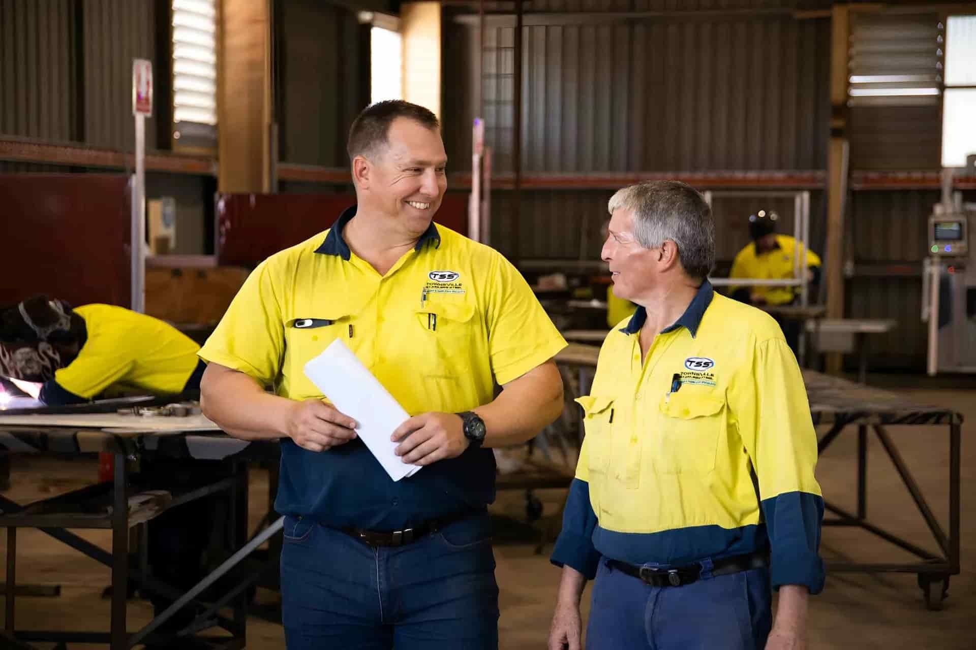 Two Workers in Yellow and Blue Work Shirts Smile at Each Other in a Workshop — Townsville Stainless Steel in Garbutt, QLD