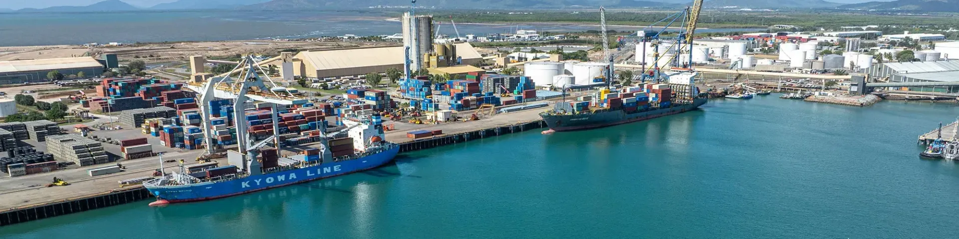 A harbor with large blue ship docked at the pier. Industrial buildings and mountains in the background.