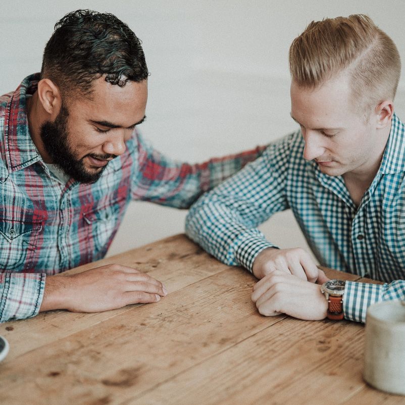 Two men are sitting at a wooden table and one has his arm around the other 's shoulder
